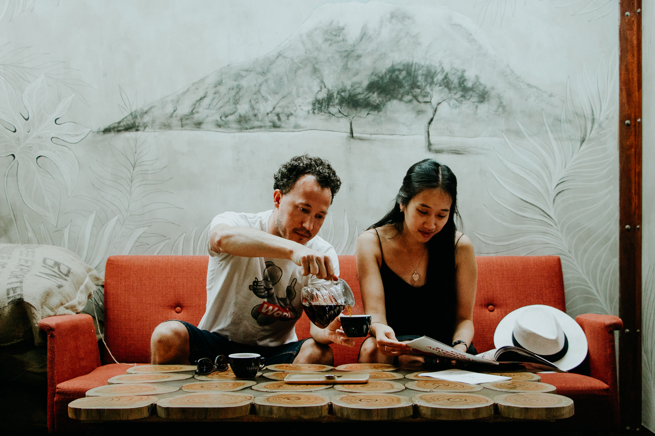 A man pours filter coffee while a woman reads a coffee magazine whilst sitting on a plush red couch inside Fabrica Coffee Roasters cafe. Lisbon, Portugal