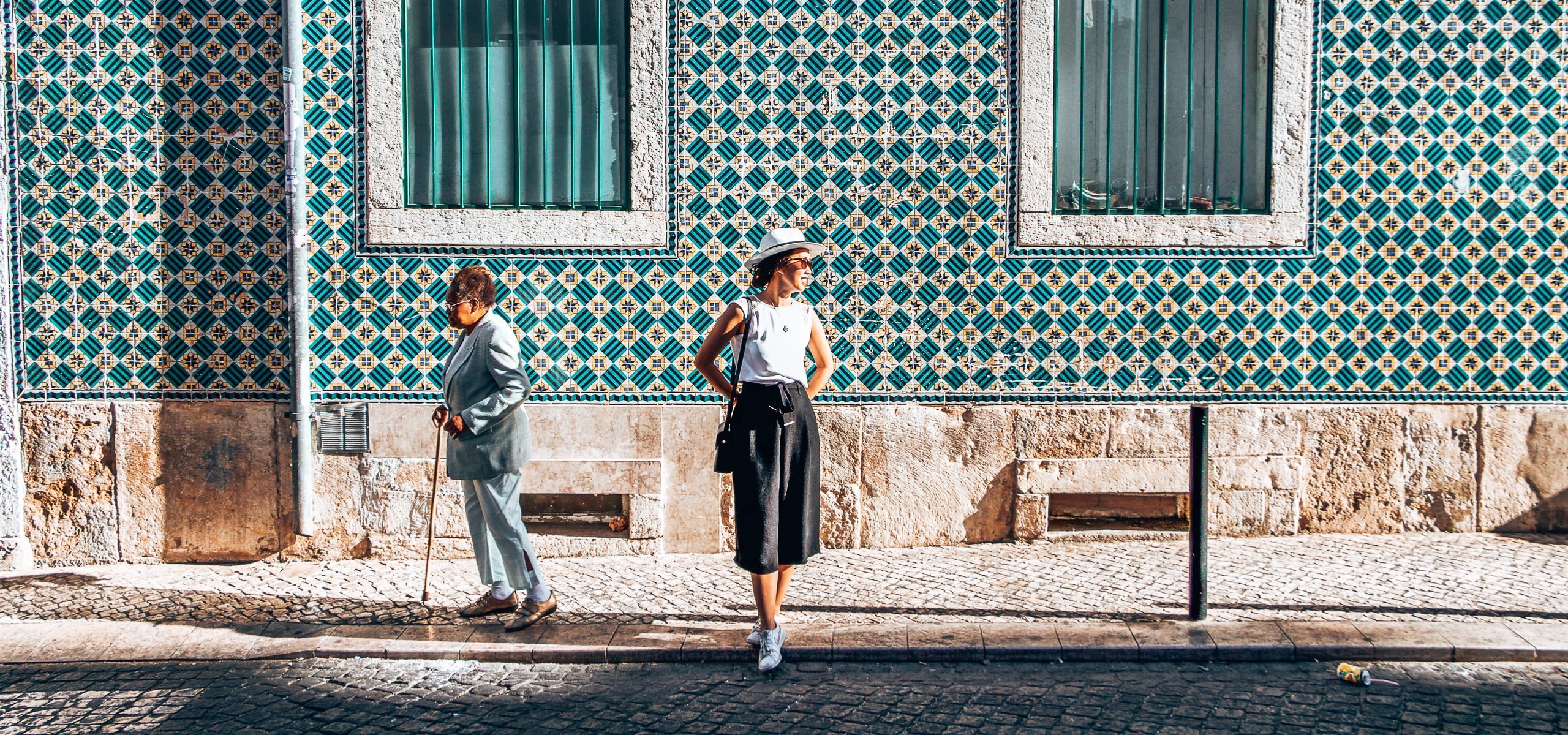 A girl stands in front of a wall bearing green and yellow azulejo tiles as an older woman with a cane shuffles past - Lisbon, Portugal