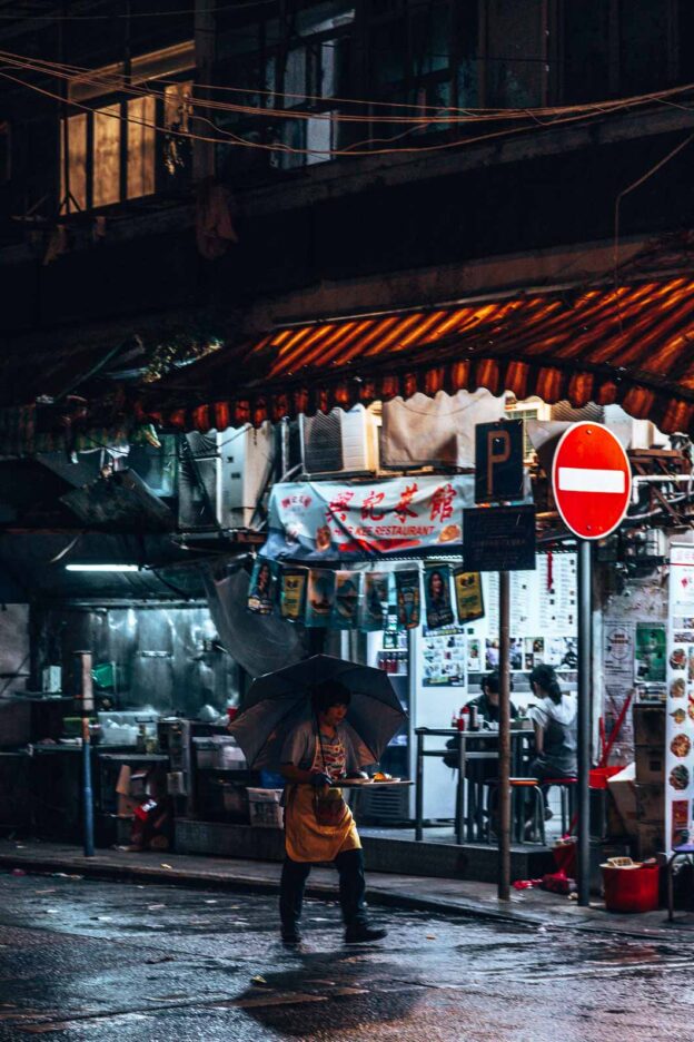 A woman holds a tray of food whilst holding am umbrella in the crook of her neck as she crosses as wet street at night in Mong Kok, Hong Kong