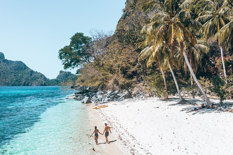 A couple walk along Ipil beach with a kayak in the background, El Nido