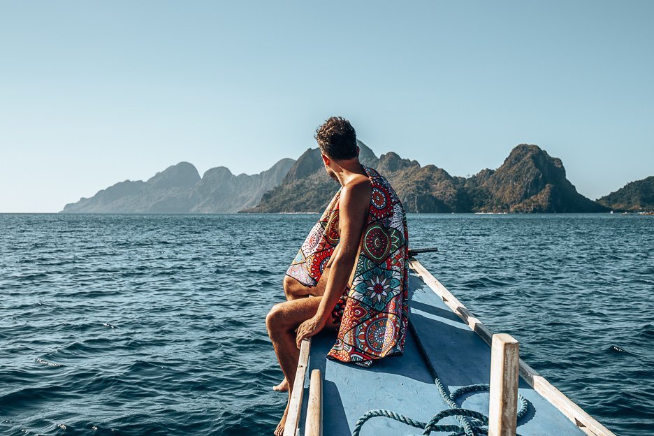 A man sits at the helm of a boat with a towel draped over his shoulders looking out towards the horizon, El Nido