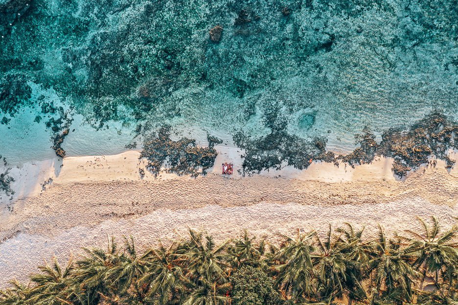 An aerial view of Cloud Nine beach. The ocean, sand and palm trees. A couple lie on a beach towel between the rocks.