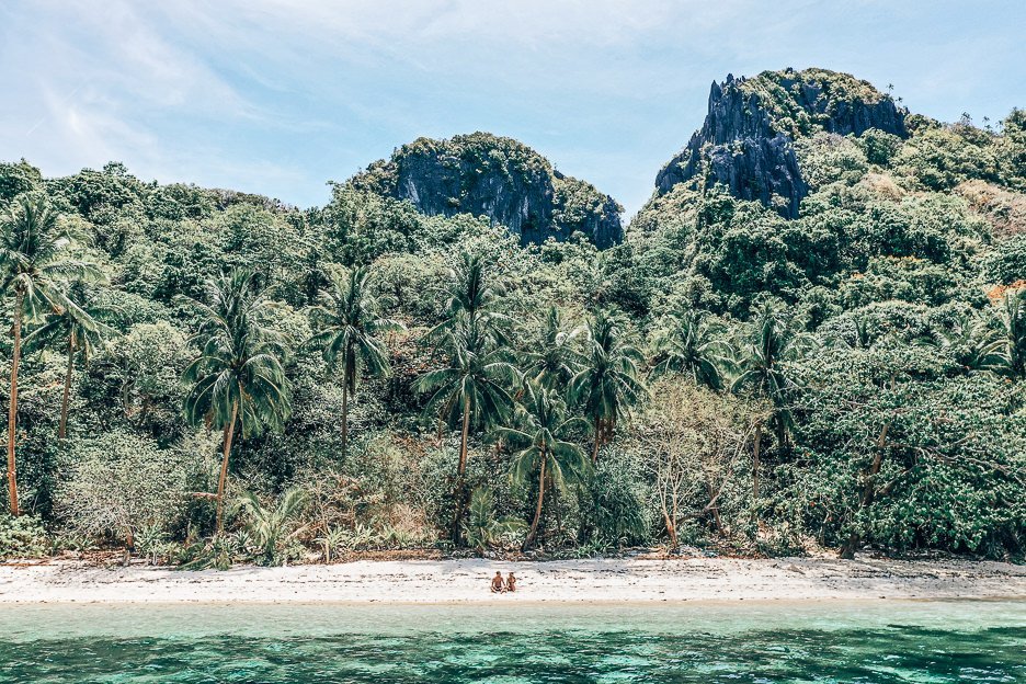 A couple sit on the pristine white sands of a tropical island paradise in El Nido