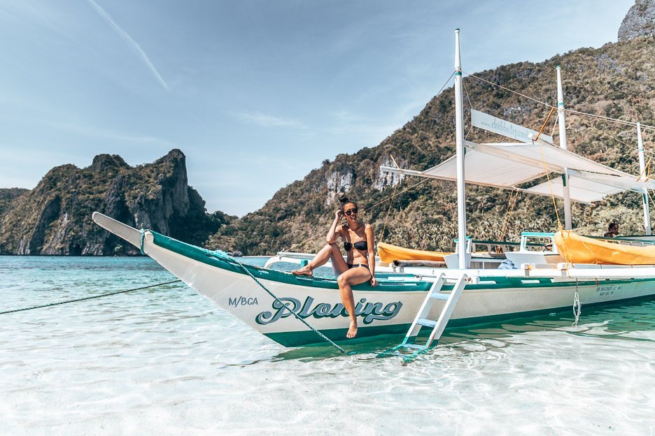 A girl in a black bikini and sunglasses sit smiling on the tour boat moored on an island in El Nido