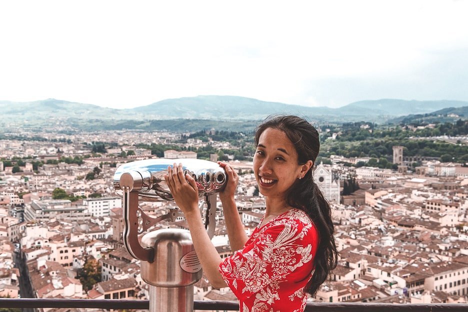 A woman in a red dress smiles at the camera whilst standing next to the binoculars at the top of the Duomo viewing platform over Florence, Italy