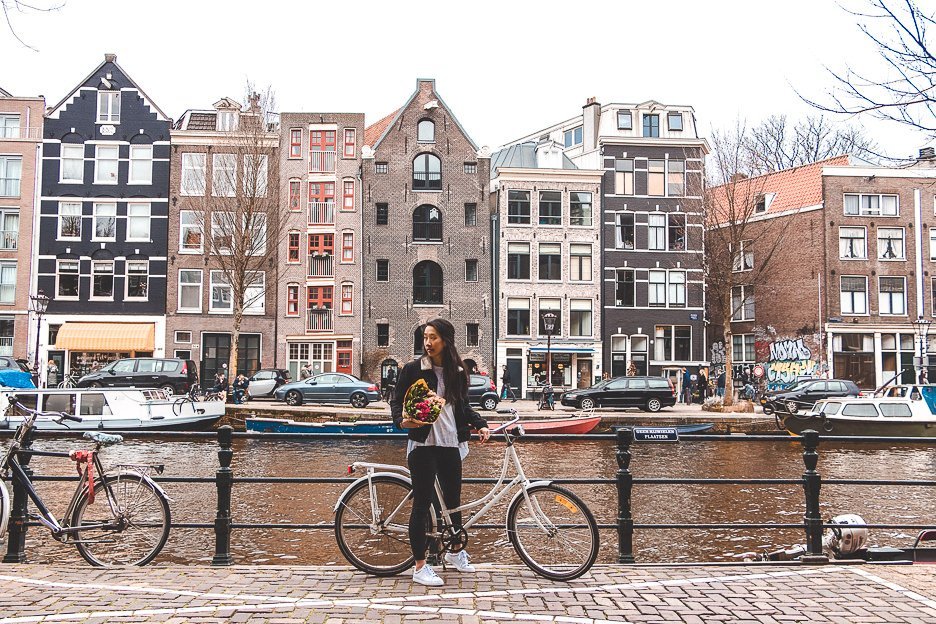 A Non Touristy Weekend Guide To Amsterdam 6 L Holding a bicycle and bunch of tulips next to the canals in Amsterdam, The Netherlands