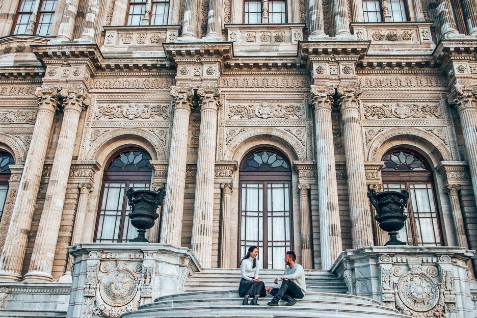 A couple sit on the stairs smiling at each other in front of Dolmabahce Palace in Istanbul, Turkey