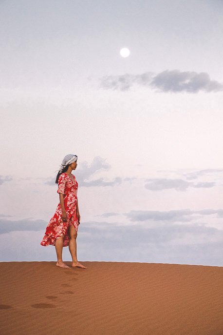 A woman in a red dress underneath the moon at dusk in the Sahara Desert, Morocco