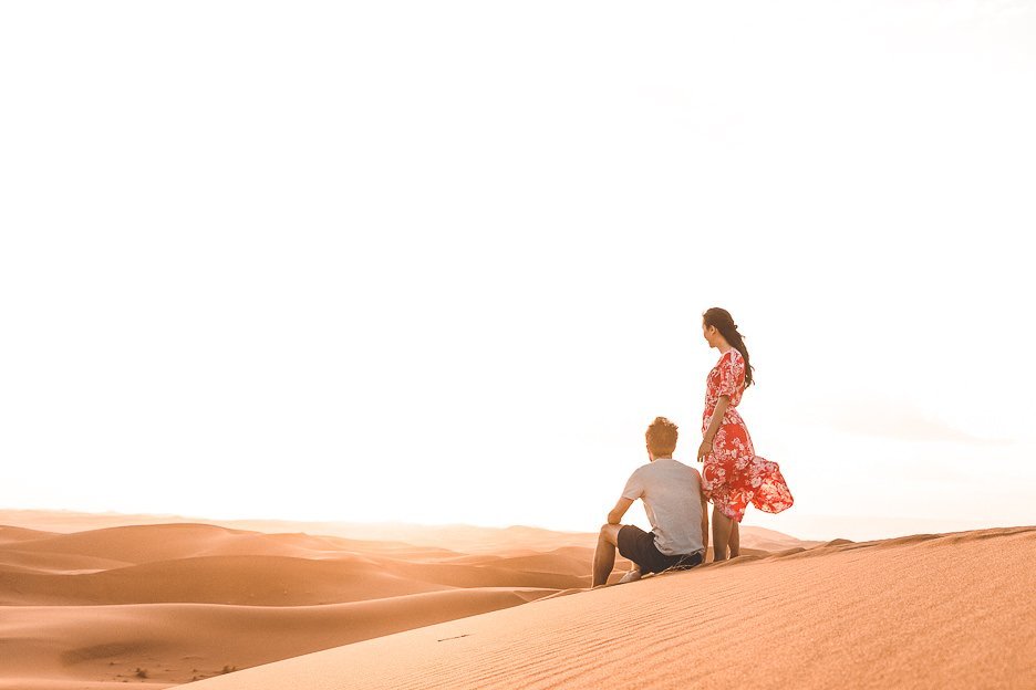 A man sits and a woman stands on a sand dune watching the sunset in the Sahara Desert, Morocco