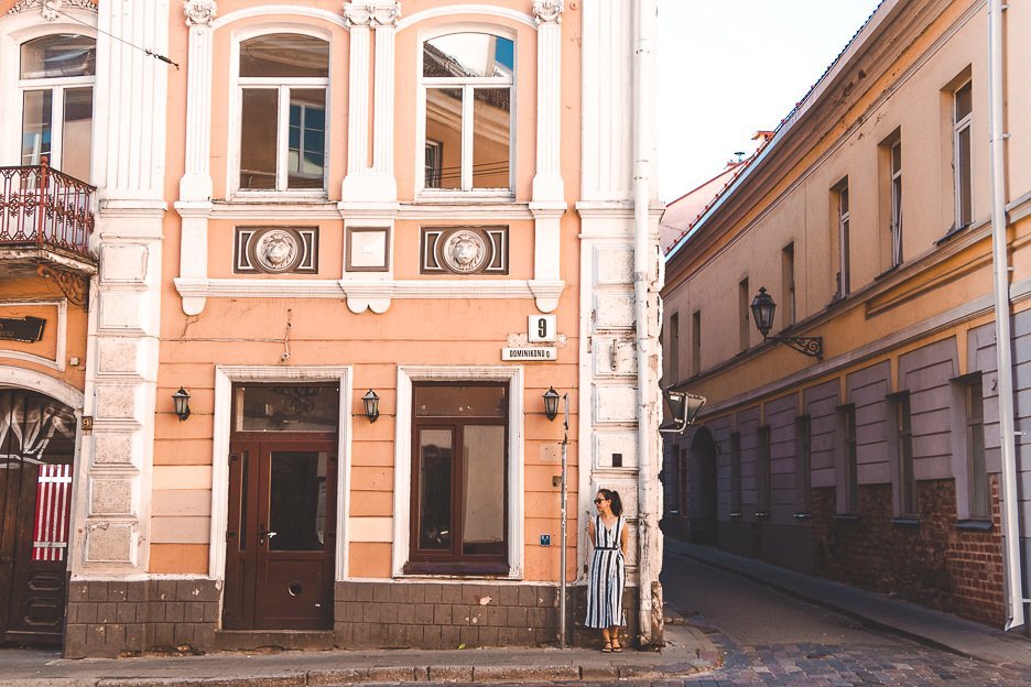 Jasmine stands in front of a pale pink building in the streets of Vilnius