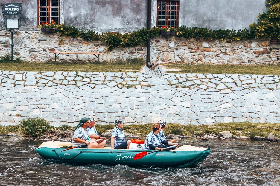 A green raft with 5 paddlers in stripey tshirts float along the Vlatva River, Český Krumlov