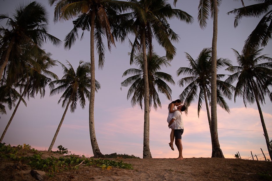 A couple share kiss underneath the palm trees on Nacpan Beach with a beautiful sunset backdrop