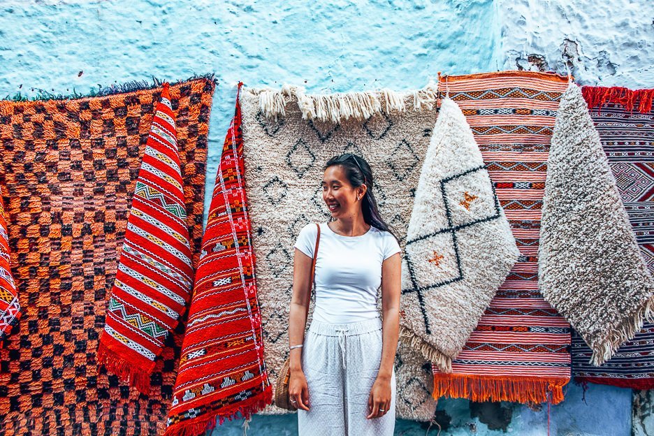 A woman wearing white stands in front of colourful woven rugs in Chefchaouen, Morocco
