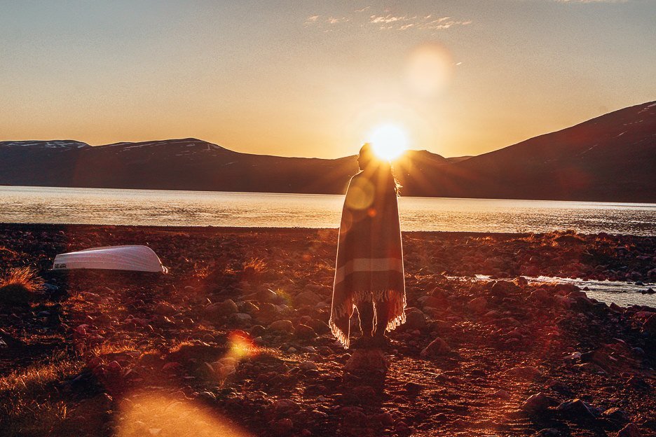 Jasmine stands on the shoreline in Swedish Lapland with a blanket wrapped around her during sunset, Sweden