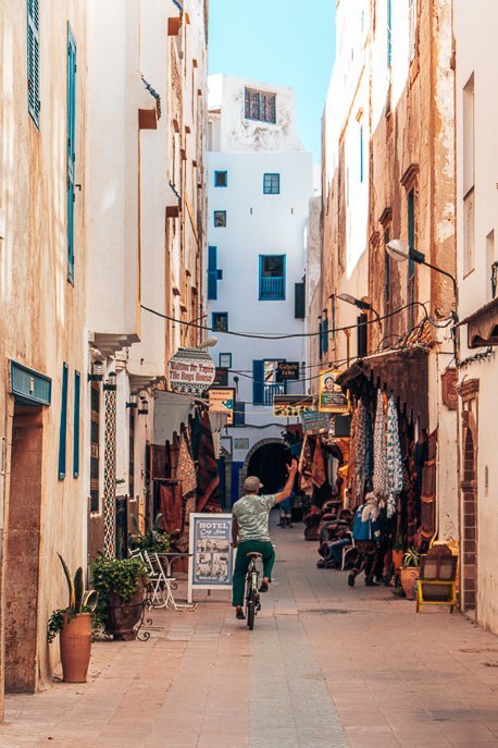 A Moroccan man rides a bicycle and waves down a street in Essaouira, Morocco