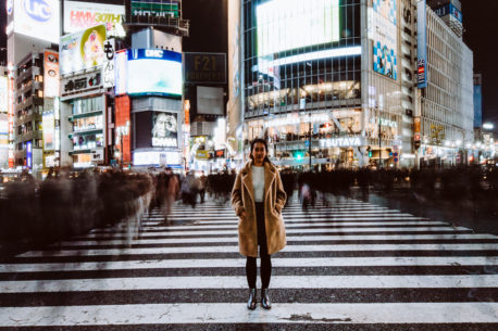 A woman stands still for a long exposure photo of Shibuya Crossing by night. Blurry pedestrians and neon lights fill the background. Tokyo, Japan