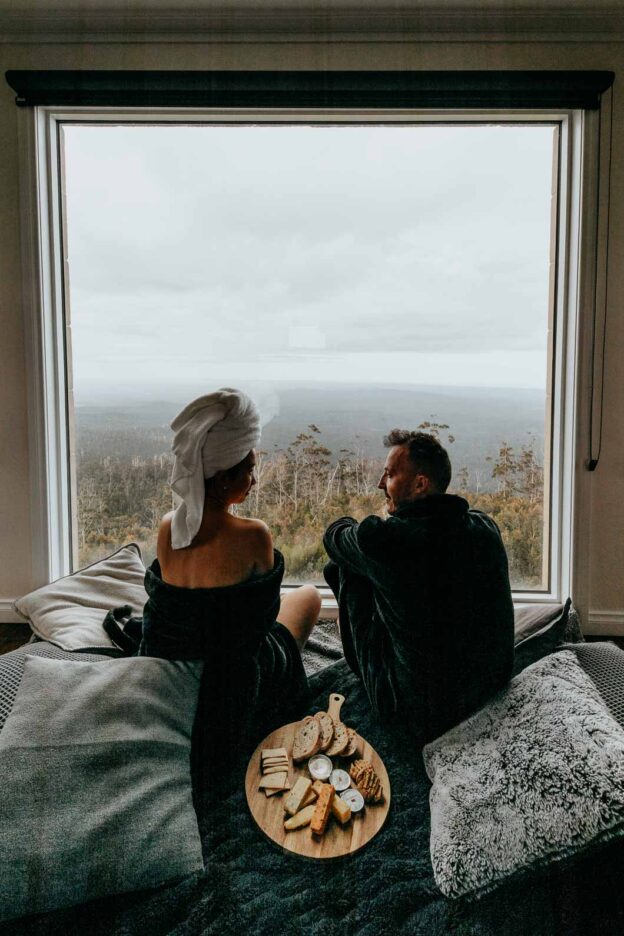 A man and woman dressed in black plush robes enjoy a cheese platter and the view from the top level of The Keep. Tasmania, Australia