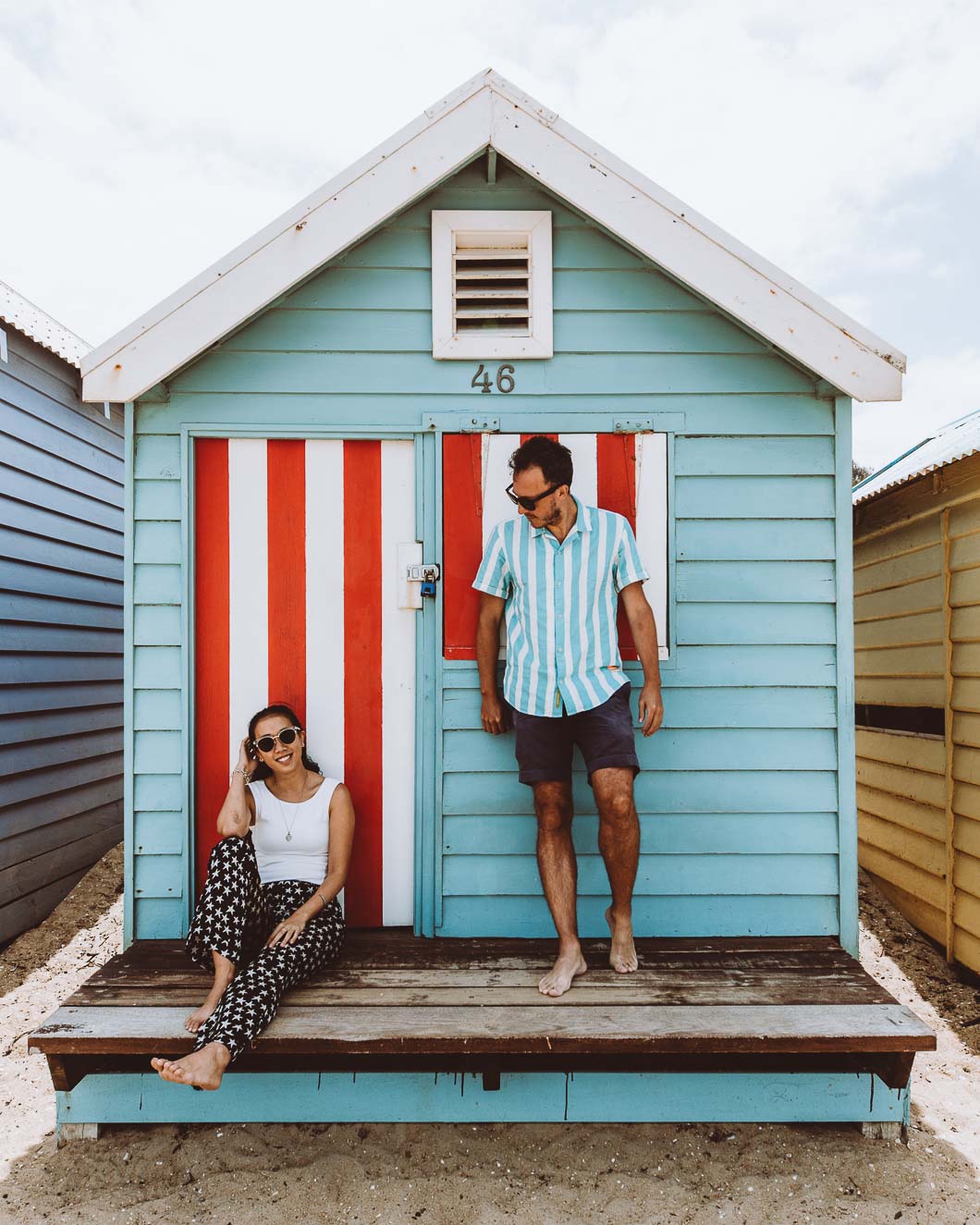 A couple on the porch of a blue bathing house in Brighton, Victoria. The door and window shade are painted red and white stripes.
