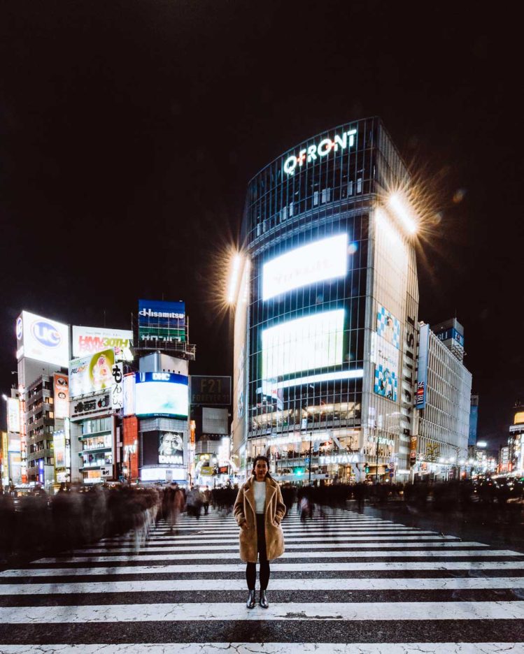 A long exposure of a woman standing motionless in Shibuya Crossing, the movement of pedestrians and the neon lights in the background. Tokyo, Japan