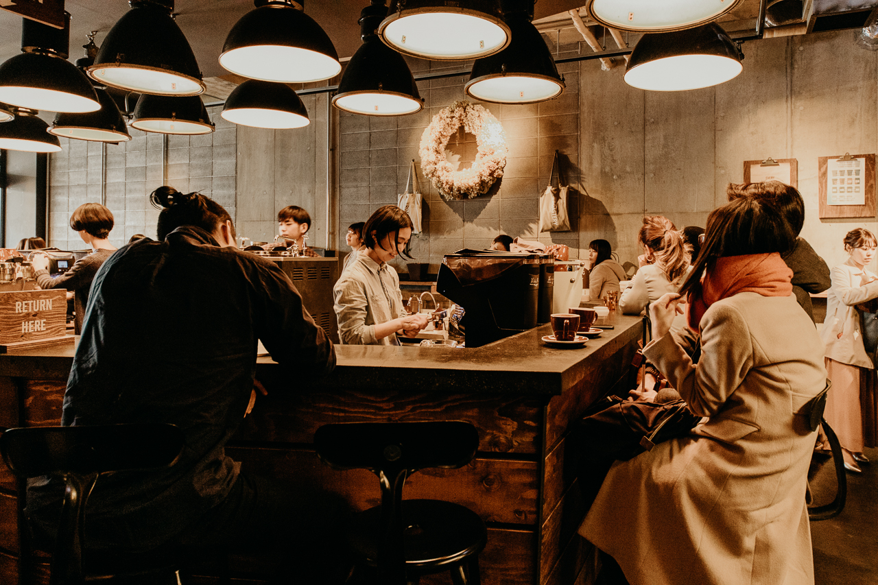Baristas prepare brews in a central station whilst customers sit on bench seats surrounding them inside The Roastery by Nozy Coffee cafe. Tokyo, Japan